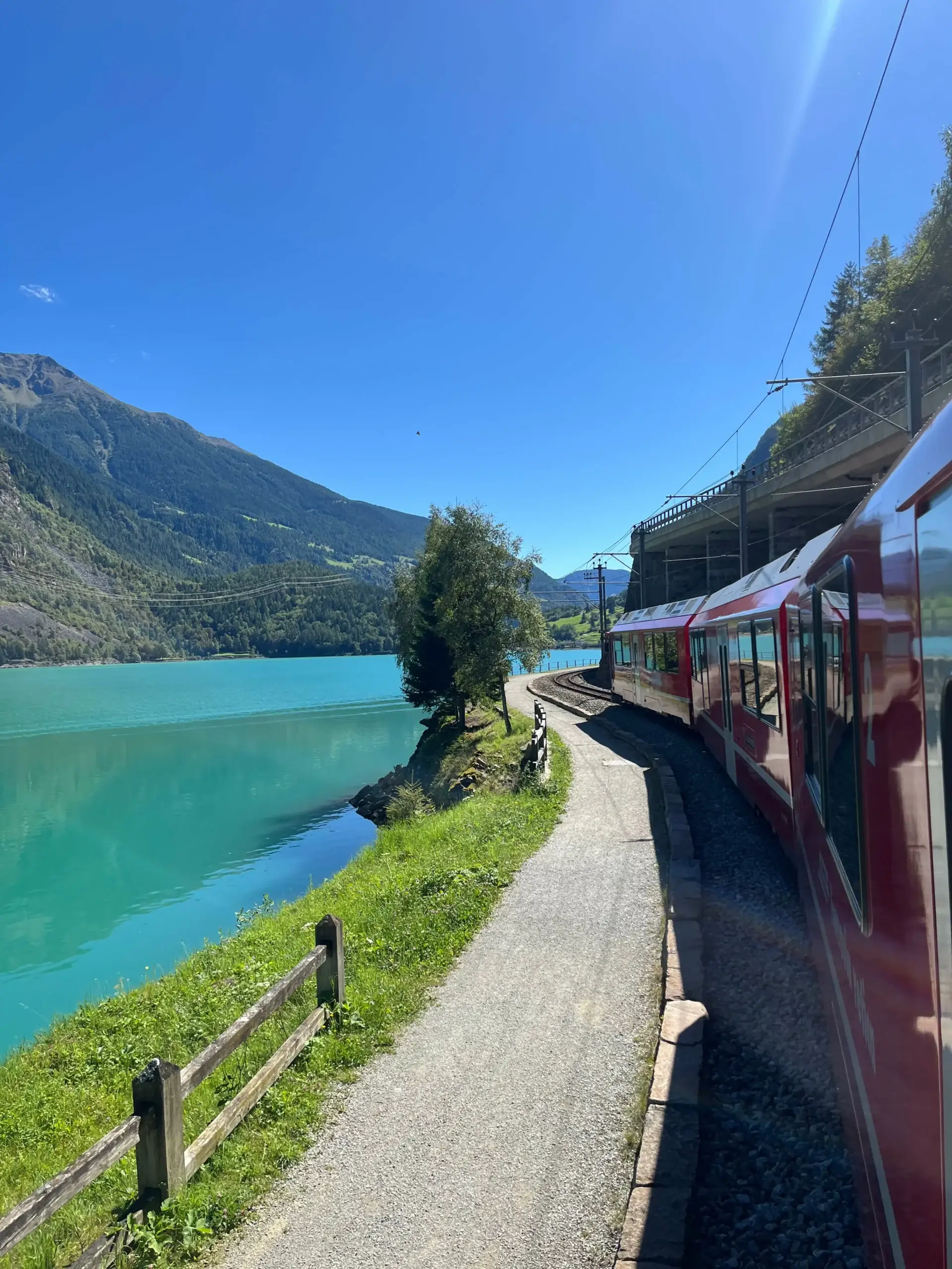 Bernina Express-toget kører langs den turkisblå Lago di Poschiavo med bjerglandskab i baggrunden under en klar blå himmel