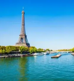 Eiffeltårnet bag grønne træer ved Seinen med flodbåde og klar blå himmel i Paris