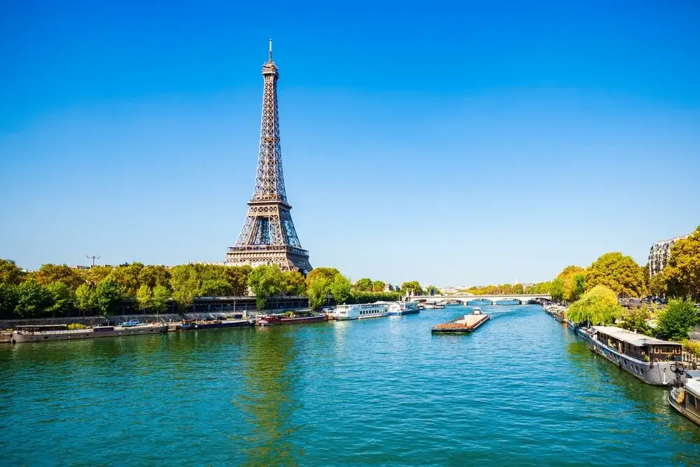 Eiffeltårnet bag grønne træer ved Seinen med flodbåde og klar blå himmel i Paris