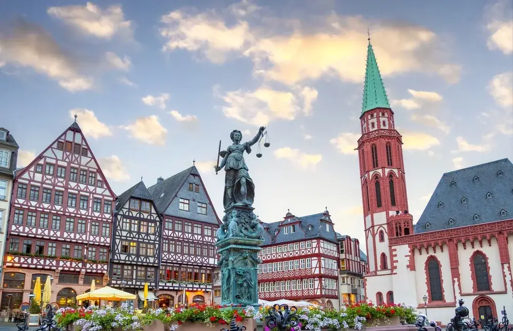 Justitia-springvandet foran bindingsværkshusene på Römerberg i Frankfurt med blomster og et rødt kirketårn