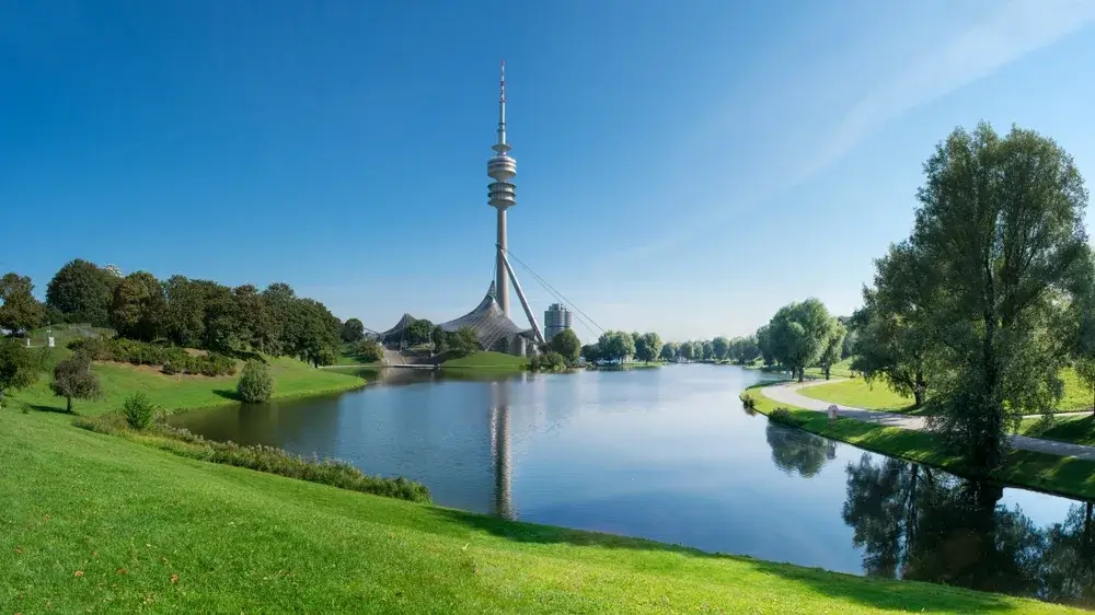 Olympiatårnet i München spejlet i søen i Olympiapark med grønne skrænter og træer om sommeren