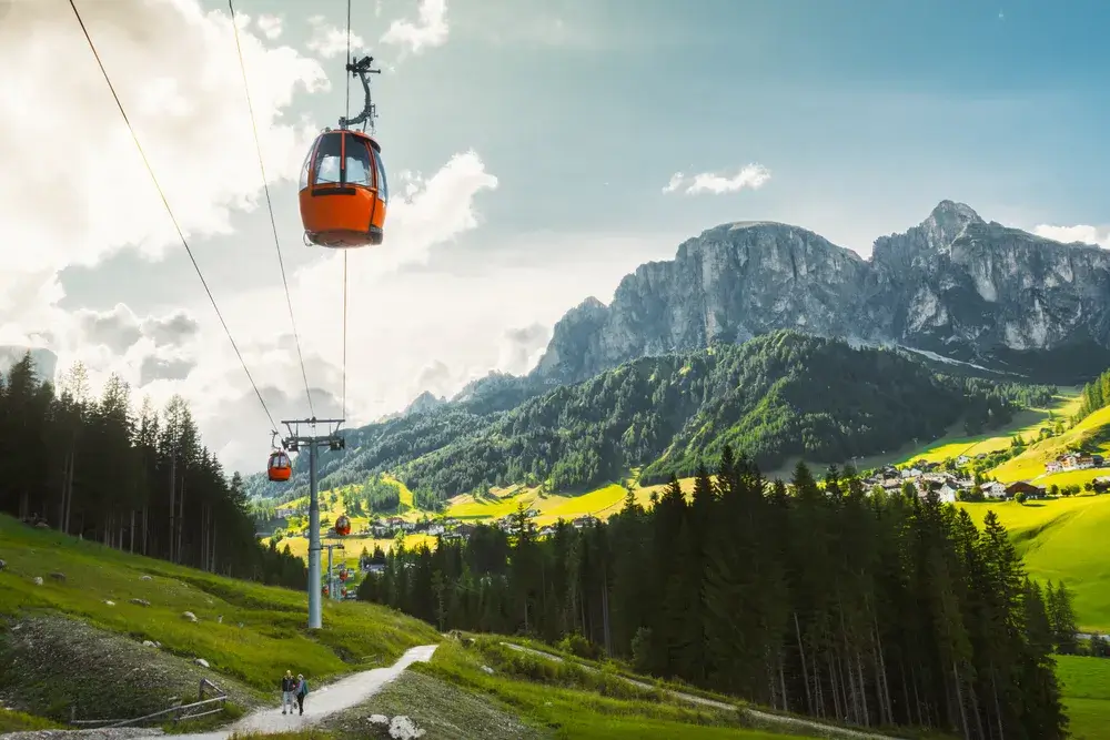 Orange gondol på kabelbane over grønne enge med Dolomitternes tinder bagved