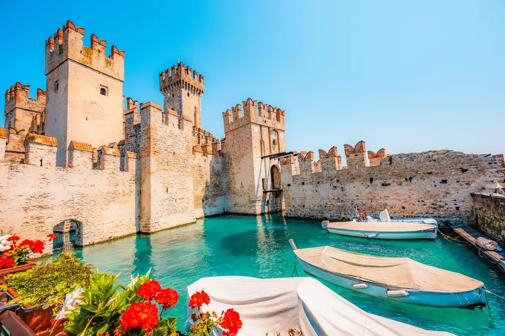 Borgen Rocca Scaligera i Sirmione omgivet af turkist vand og røde blomster i forgrunden