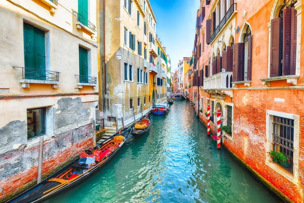 Turkis kanal mellem farverige facader i Venedig med en fortøjet gondol og stribede fortøjningspæle
