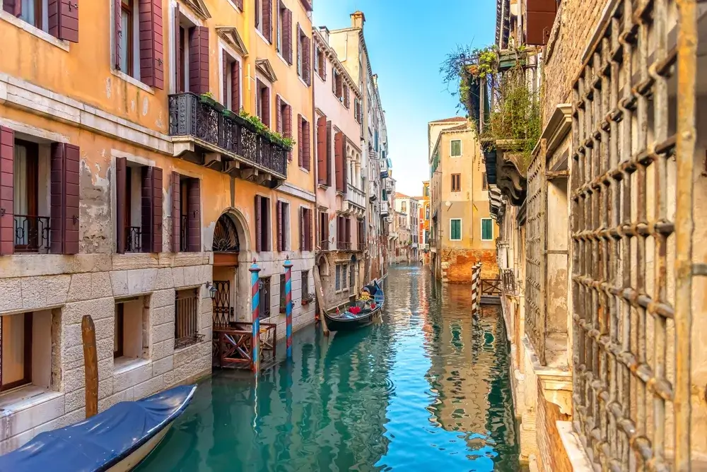 Smal kanal i Venedig mellem gamle okkerfarvede huse med altaner og en gondol i baggrunden