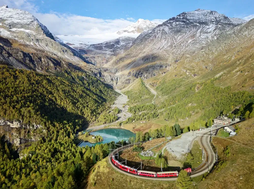 Rødt tog i kurven under Alp Grüm på Bernina-banen med Lago Palü og bjergene over Valposchiavo