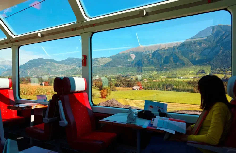 Passager ved bordet i en panoramavogn med store vinduer ud mod grønne enge og bjerge i Schweiz