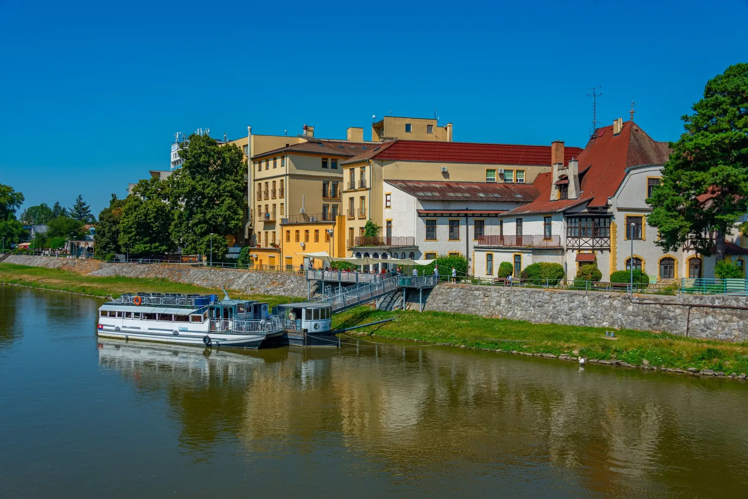 Floden Váh med en flodbåd ved kajen og byens huse i Piešťany