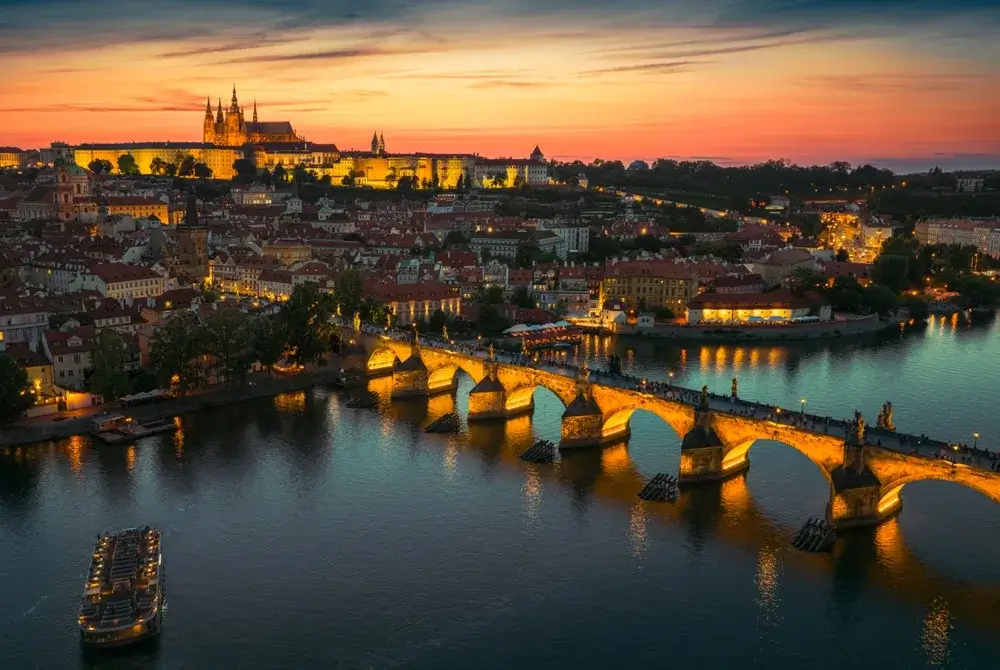 Luftfoto af Prag ved solnedgang med den oplyste Karlsbro over Vltava og Prag Borg i baggrunden