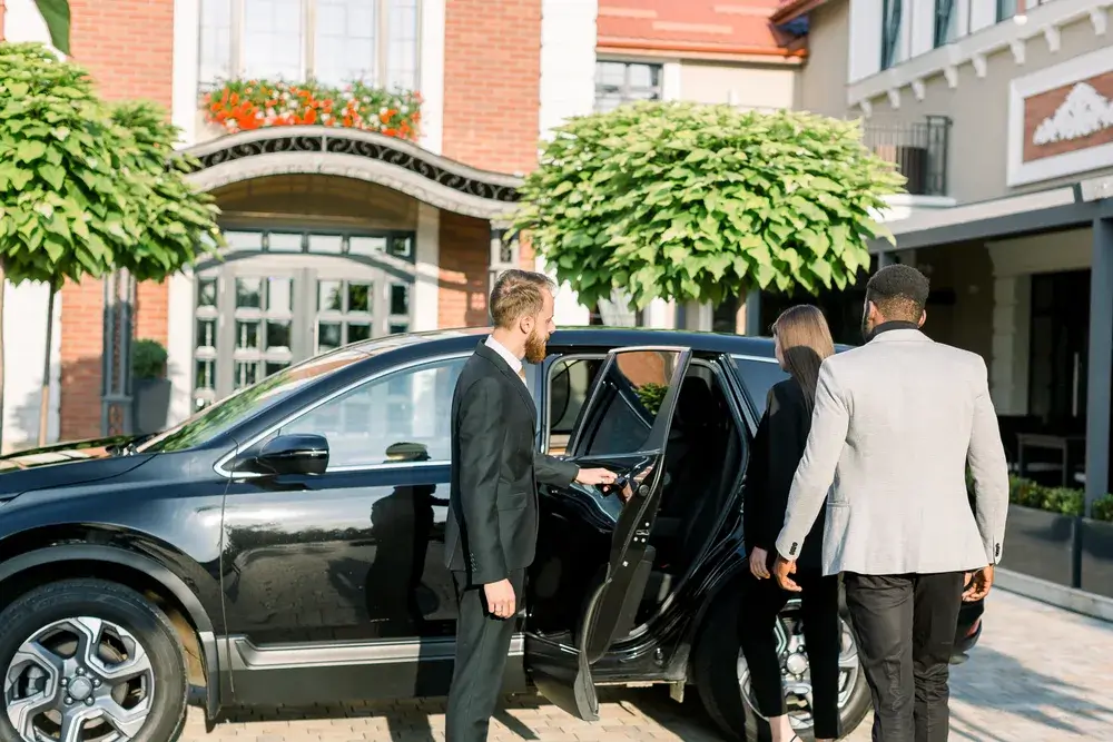 Chauffør i mørkt jakkesæt åbner bagdøren på en sort bil for to rejsende foran et hotel